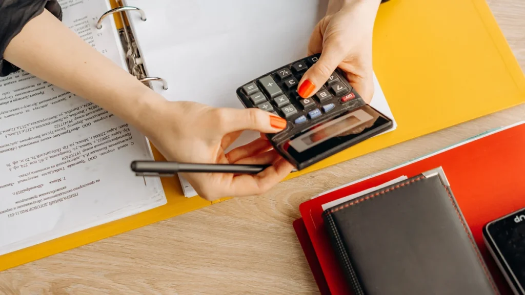 Hands using a calculator beside financial paperwork and folders on a desk.