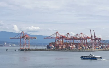 Container cranes and cargo stacks at a Vancouver port terminal, with mountains in the background.
