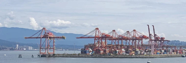 Container cranes and cargo stacks at a Vancouver port terminal, with mountains in the background.