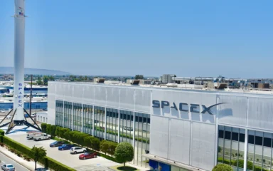 SpaceX factory building in Hawthorne, California, with the SpaceX logo and a Falcon 9 booster outside.