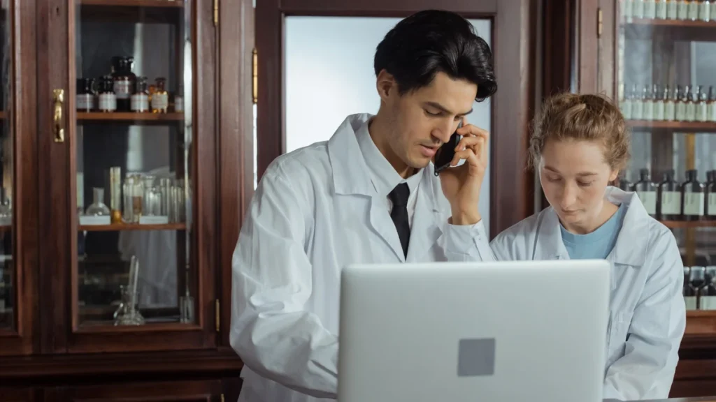 Two pharmacists in white coats using a laptop and phone behind a pharmacy counter.