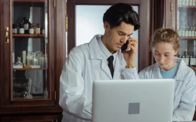Two pharmacists in white coats using a laptop and phone behind a pharmacy counter.
