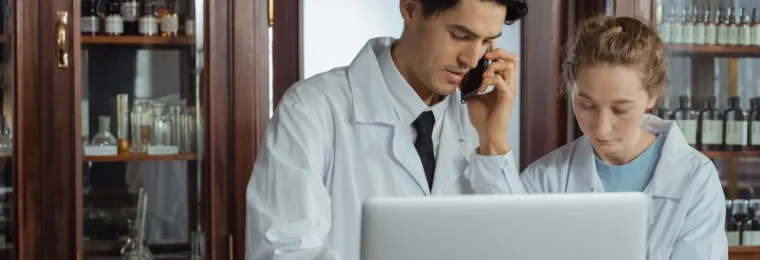 Two pharmacists in white coats using a laptop and phone behind a pharmacy counter.