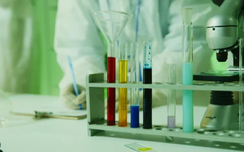 Colored liquids in laboratory test tubes beside a microscope, with a researcher in the background.