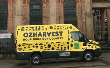 A yellow OzHarvest food delivery van parked outside St James' Church in Sydney, Australia.