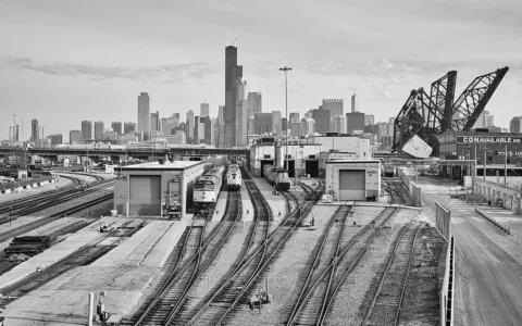 Black-and-white view of Chicago rail yards, trains, a raised rail bridge and the downtown skyline.
