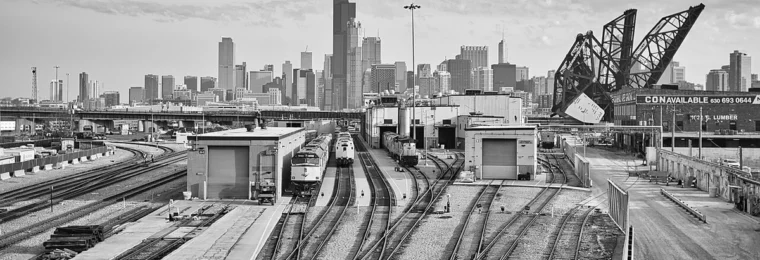 Black-and-white view of Chicago rail yards, trains, a raised rail bridge and the downtown skyline.