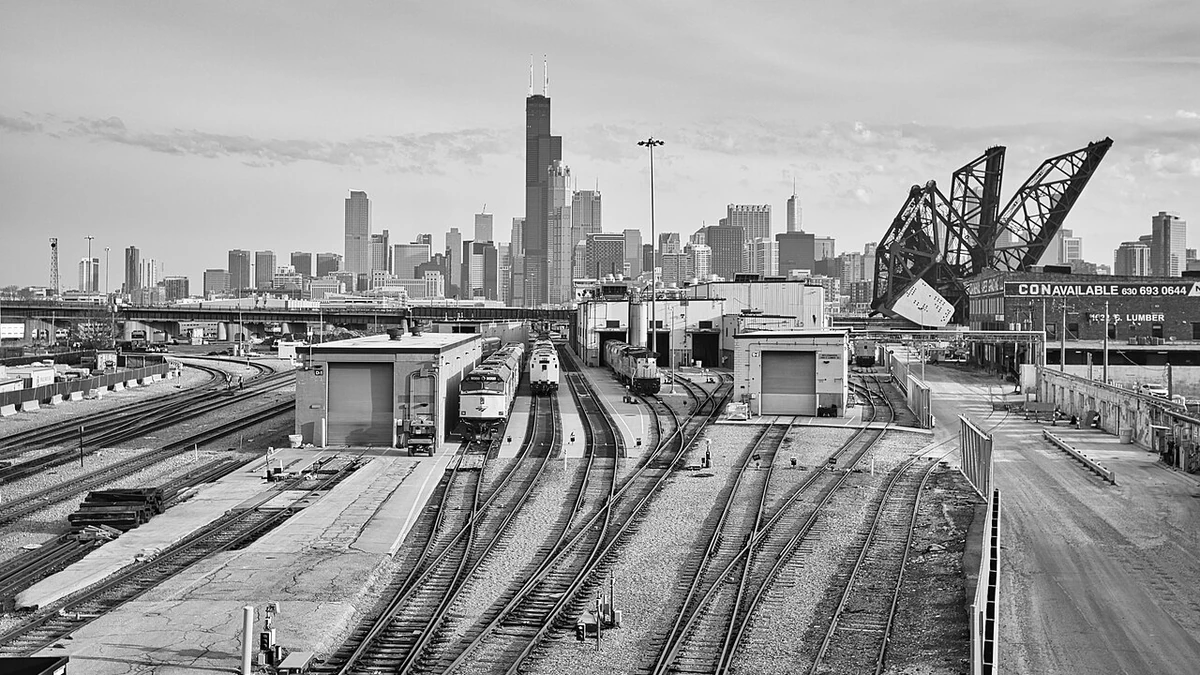 Black-and-white view of Chicago rail yards, trains, a raised rail bridge and the downtown skyline.