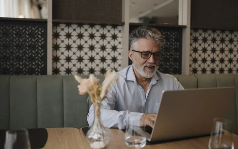 An older man with gray hair and glasses using a laptop at a cafe table.