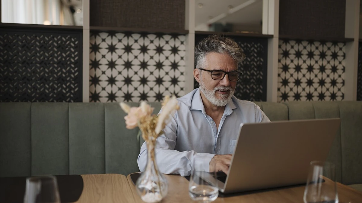An older man with gray hair and glasses using a laptop at a cafe table.