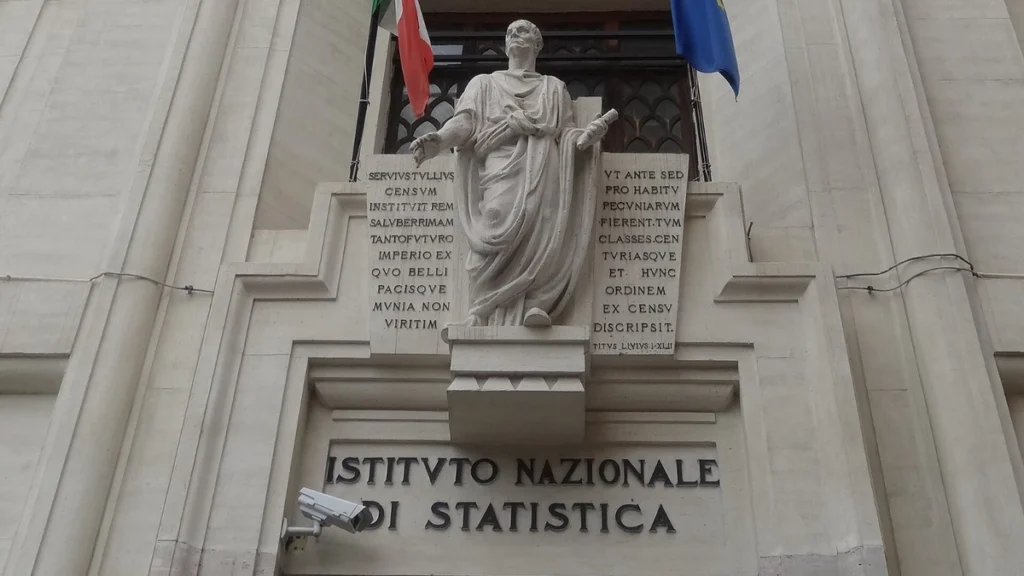 Entrance of the Istituto Nazionale di Statistica building in Rome with the Istat signage visible below a statue and Italian and European Union flags.