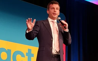 David Seymour speaking into a microphone in front of ACT Party signage at a 2023 party meeting in Palmerston North.
