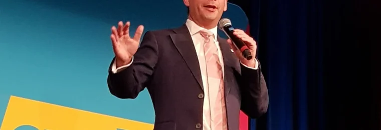 David Seymour speaking into a microphone in front of ACT Party signage at a 2023 party meeting in Palmerston North.
