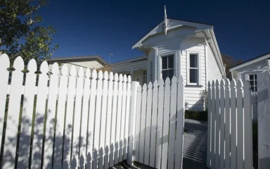 A white wooden house with a picket fence in Northcote Point, Auckland, under a blue sky.