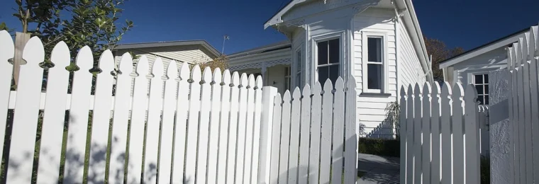 A white wooden house with a picket fence in Northcote Point, Auckland, under a blue sky.