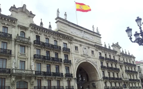 Stone facade of a Banco Santander building in Santander, Spain, with a Spanish flag flying over the central arch.