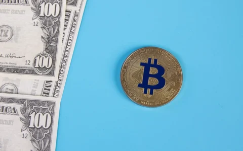 A gold-colored Bitcoin token on a blue background beside U.S. $100 bills.