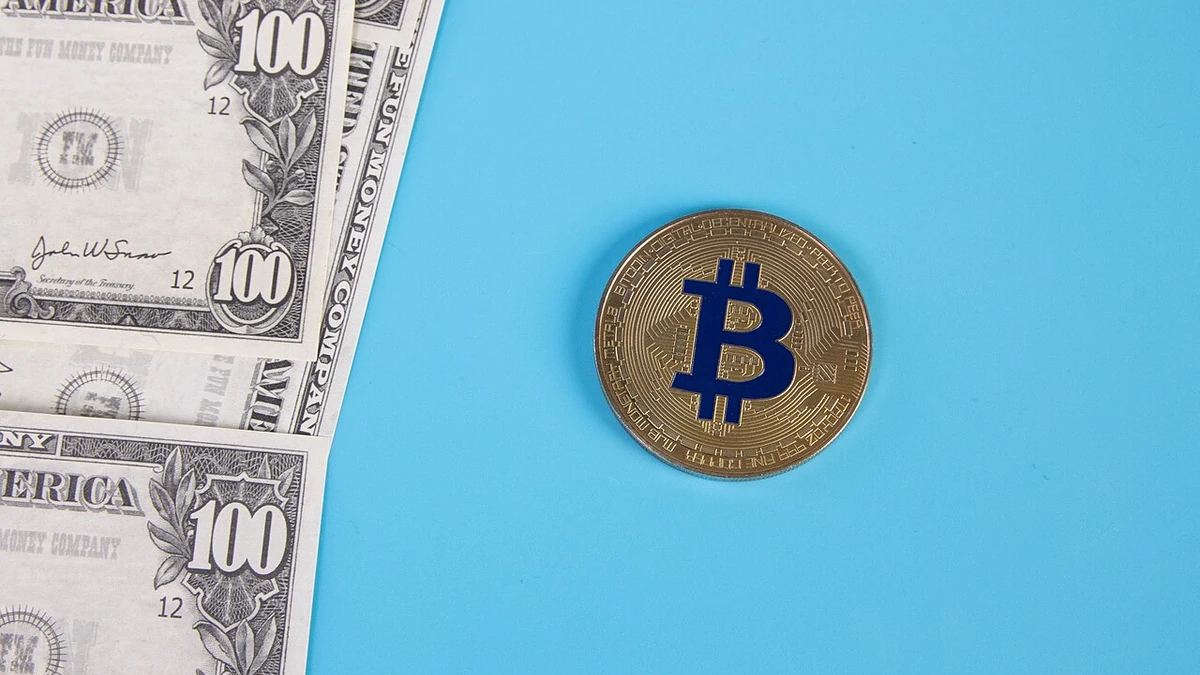 A gold-colored Bitcoin token on a blue background beside U.S. $100 bills.