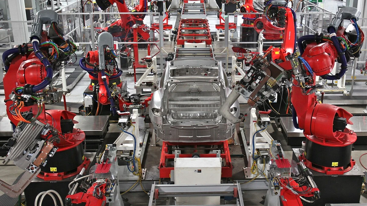 Red industrial robots surround an unfinished car body on a Tesla Model S assembly line.