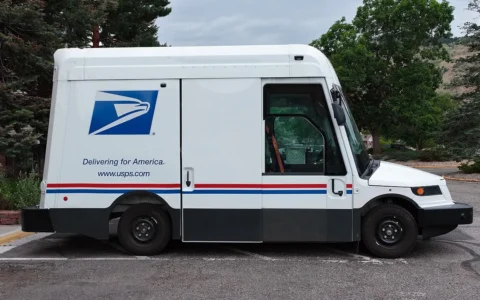 Side view of a white USPS Oshkosh Next Generation Delivery Vehicle with the Postal Service logo.