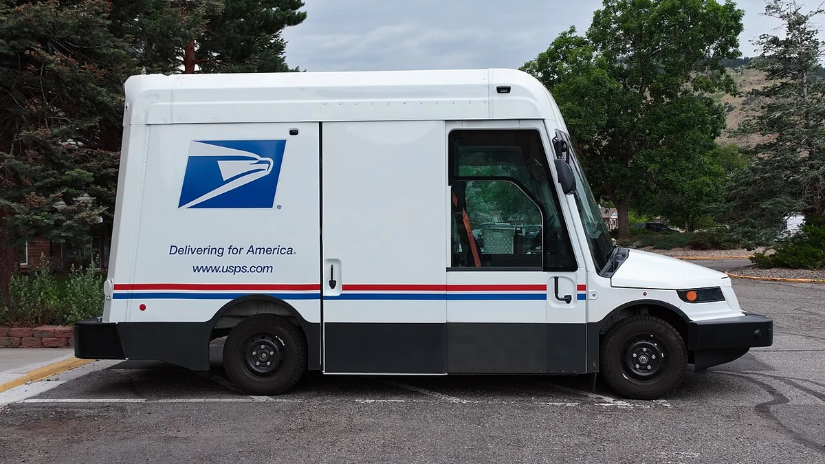 Side view of a white USPS Oshkosh Next Generation Delivery Vehicle with the Postal Service logo.