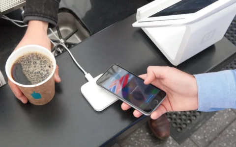 A customer holds an iPhone beside a Square contactless payment reader while buying coffee.